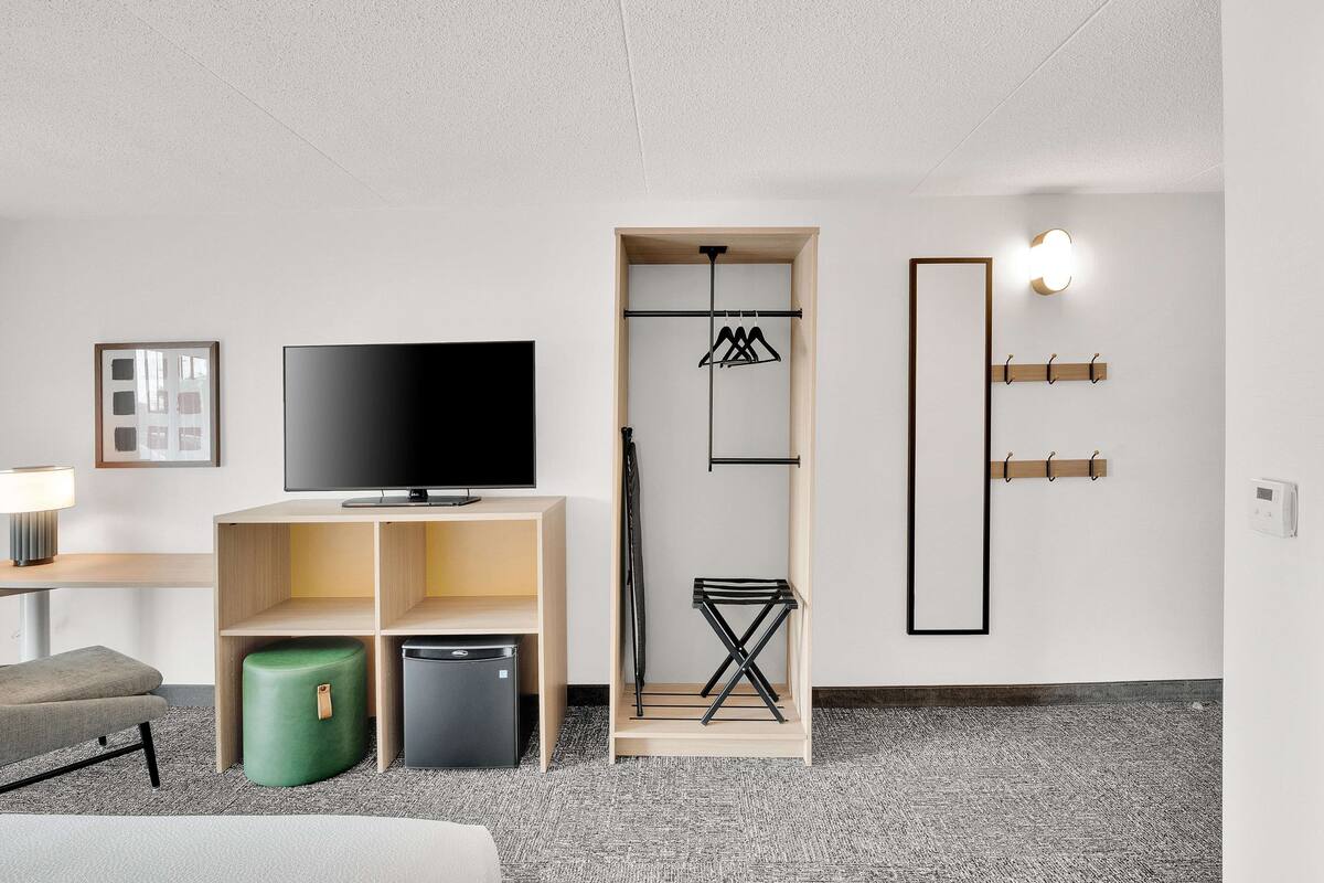 A modern hotel room with a flat-screen TV, a wooden TV stand, a green ottoman, a compact fridge, a built-in wardrobe with a hanging rack, and a wall-mounted light fixture.