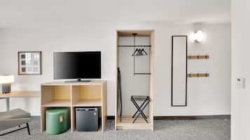 A modern hotel room with a flat-screen TV, a wooden TV stand, a green ottoman, a compact fridge, a built-in wardrobe with a hanging rack, and a wall-mounted light fixture.