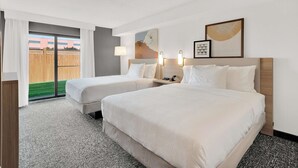 A hotel room with two beds, a large window with curtains, a view of a wooden fence, and a wall with framed artwork and wall sconces.