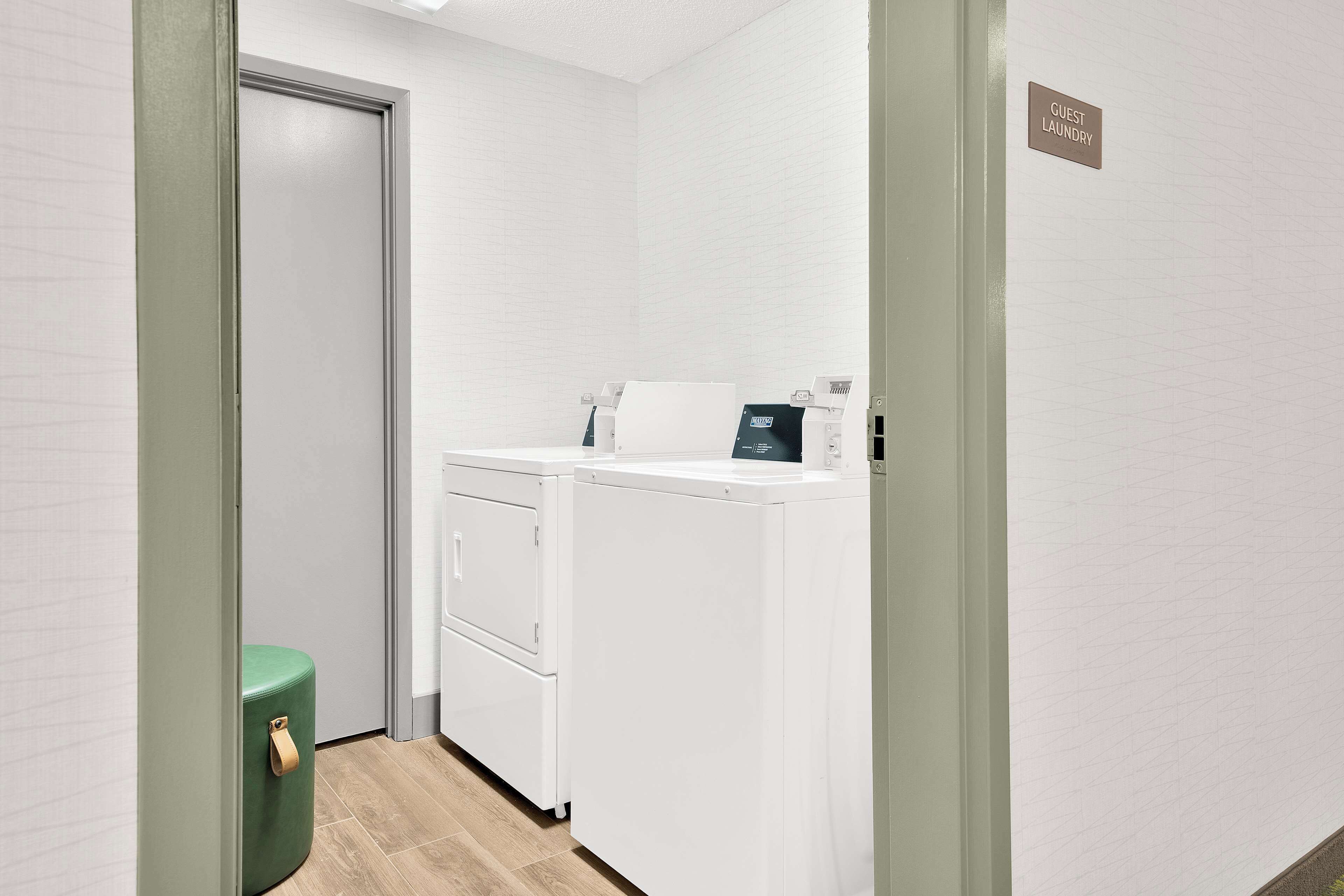 A laundry area with a green trash bin, a white washing machine, and a sign indicating guest laundry facilities.