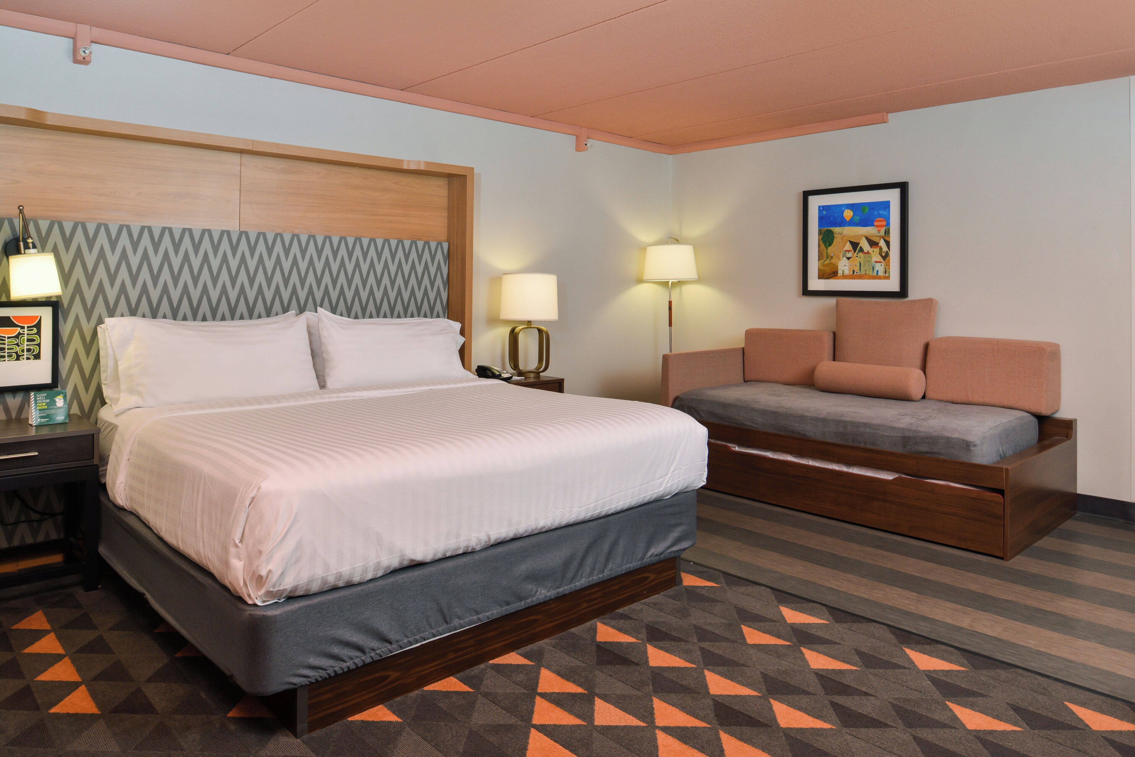 A hotel room with a bed, bedside table, lamp, and a sofa.