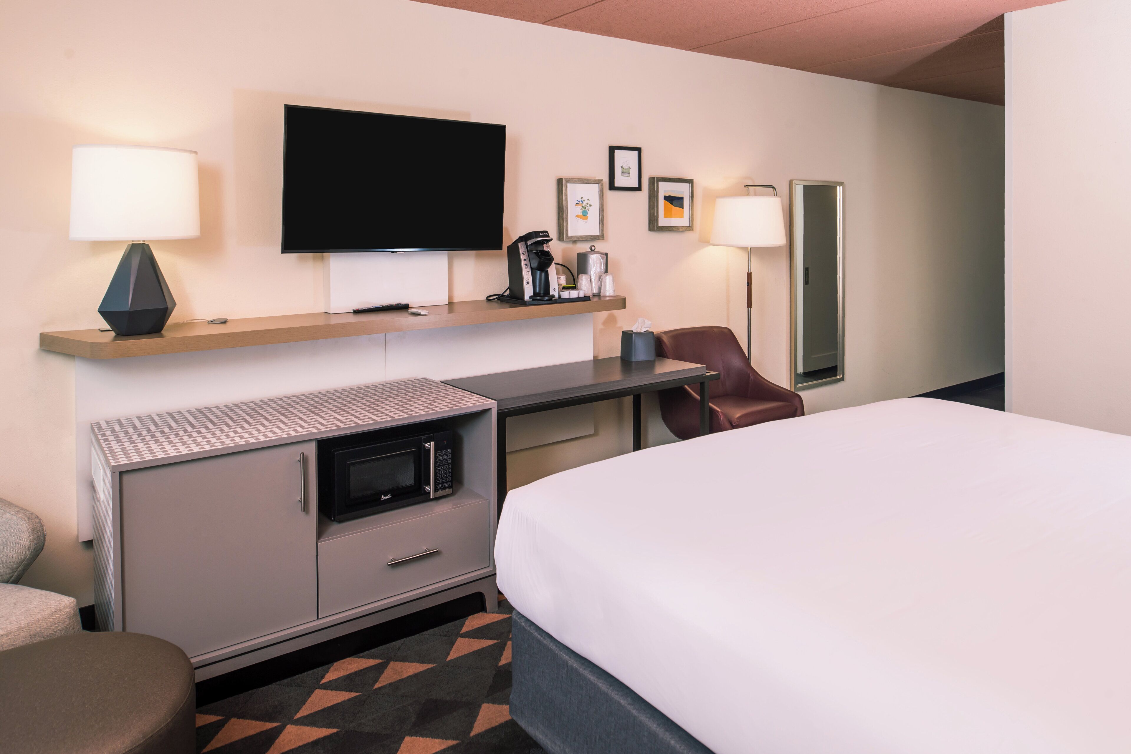 A hotel room with a bed, a desk with a flat-screen TV, a coffee maker, a microwave, and a chair.