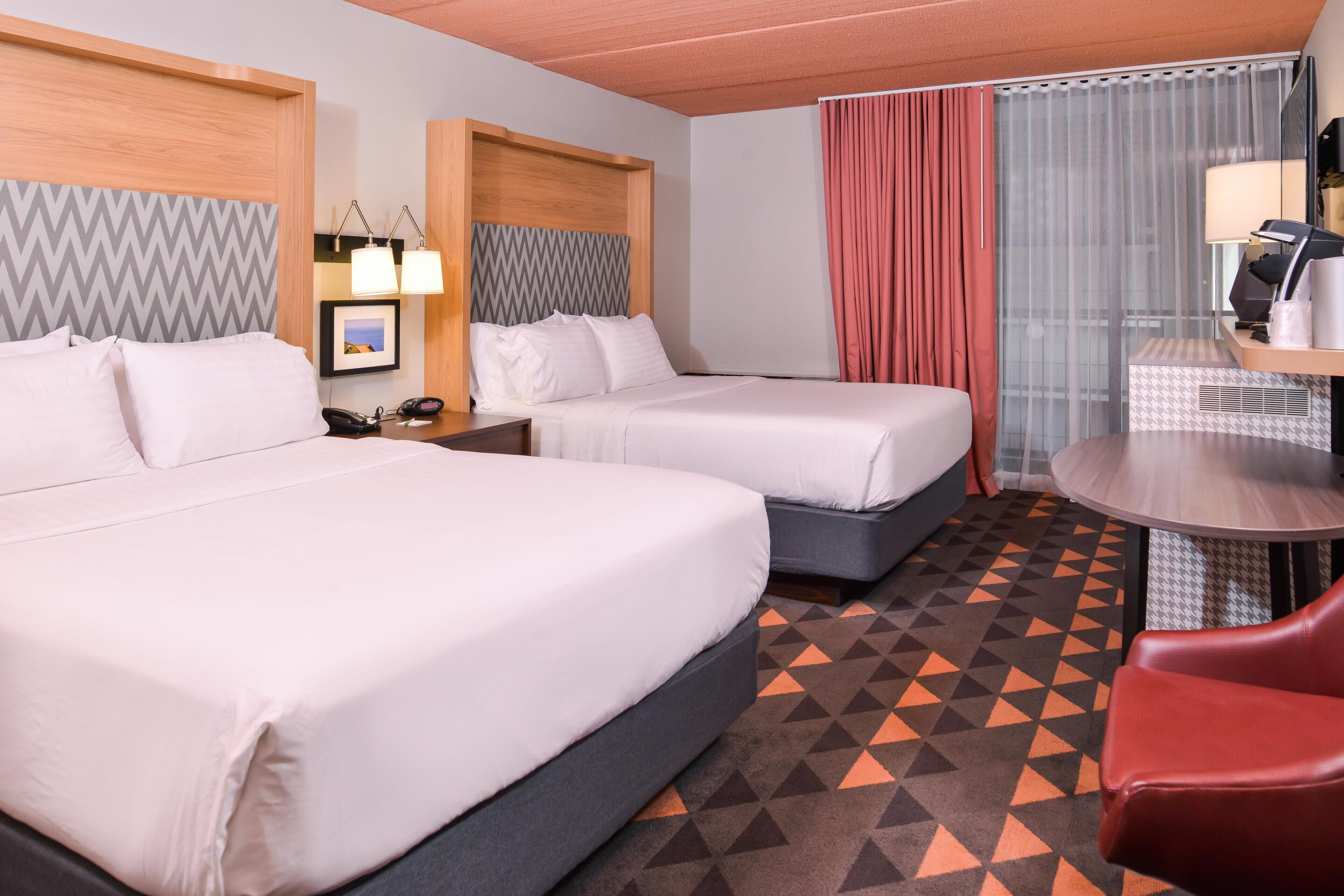 Une chambre d’hôtel avec deux lits, un bureau avec une chaise, un miroir, une lampe et une moquette à motifs.