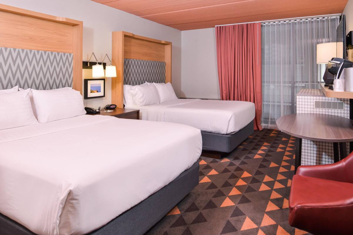 Quarto de hotel com duas camas, uma escrivaninha com cadeira, espelho, abajur e tapete estampado.