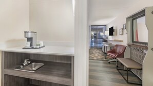 Une chambre d’hôtel équipée d’une kitchenette, d’un évier, d’une banquette, d’un miroir et donnant sur le couloir.