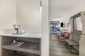 A hotel room with a kitchenette, a sink, a bench, a mirror, and a view of the hallway.