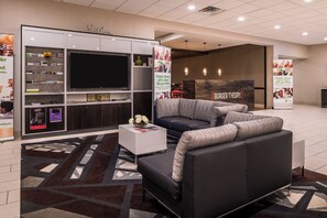 A modern hotel lobby with a large flat-screen TV, a sofa, a coffee table, and a patterned rug. - Holiday Inn Auburn-Finger Lakes Region by IHG (Auburn)