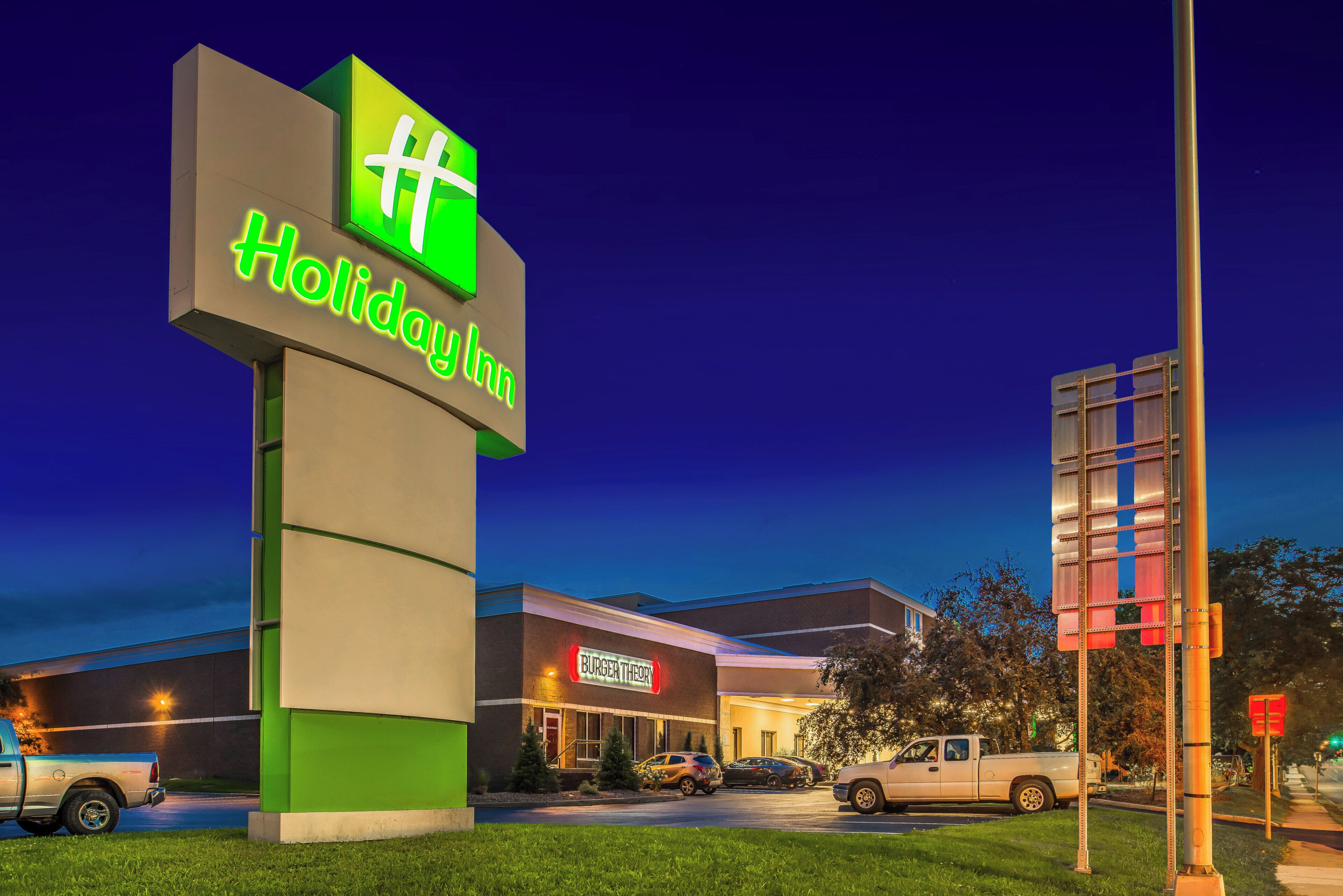 A Holiday Inn sign with a green and white logo, a brick building with a Burger Theory restaurant, and a parking lot with vehicles.