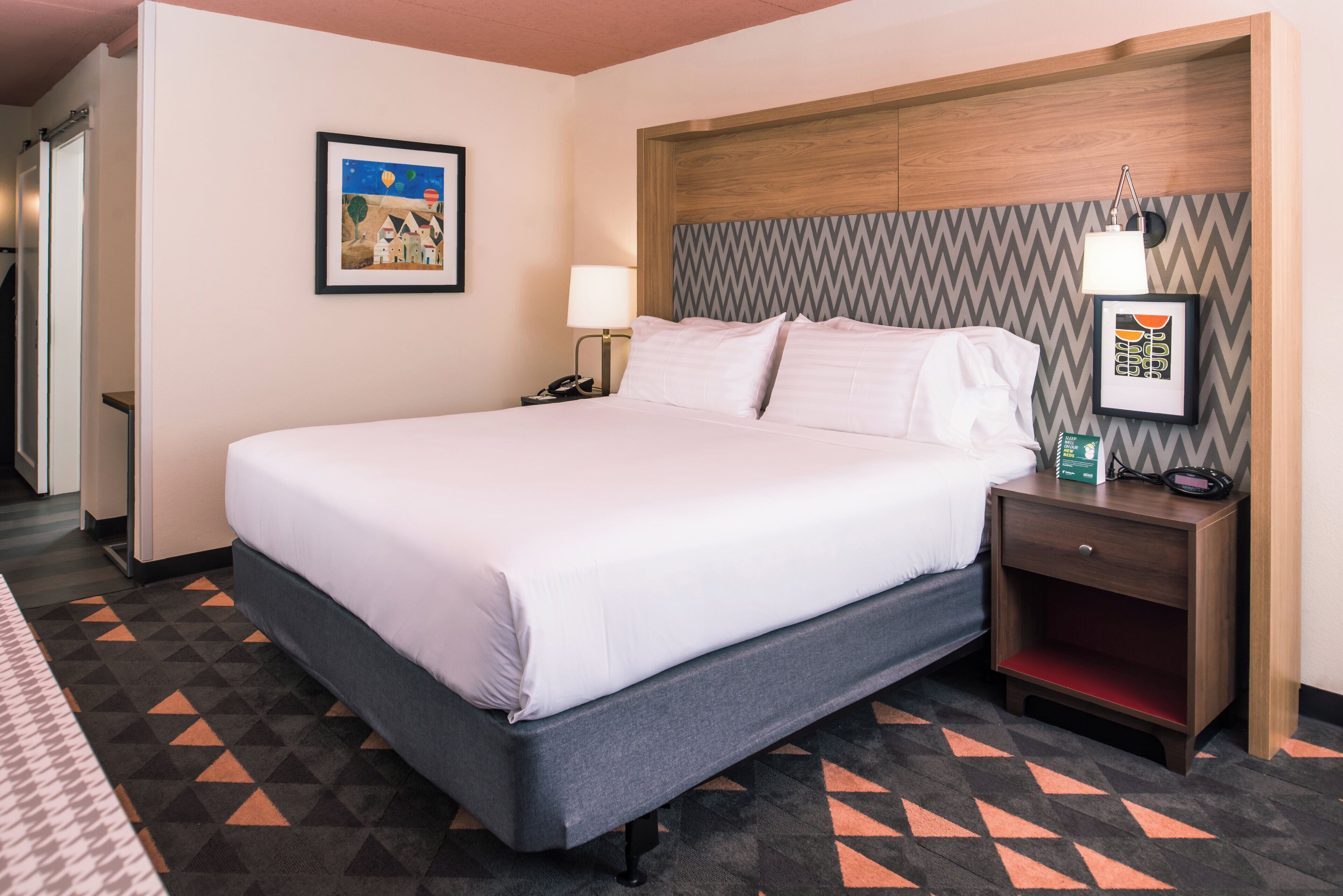 Une chambre d’hôtel avec un lit, une table de chevet, une lampe, un tableau encadré au mur et une moquette à motifs.