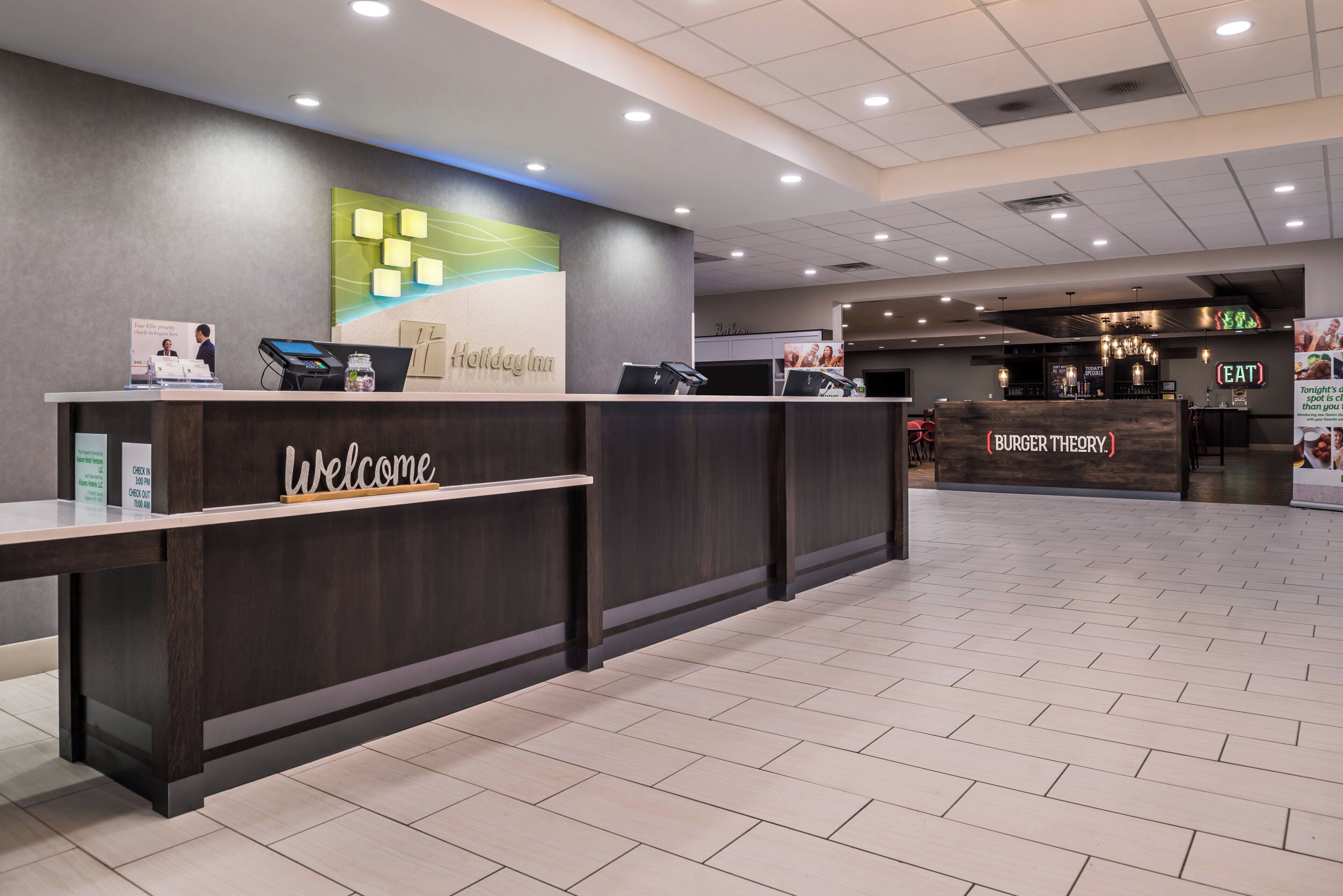 Un hall d’accueil d’hôtel avec un comptoir de réception en bois foncé, une pancarte « Bienvenue » et une enseigne « Holiday Inn » au-dessus du comptoir.