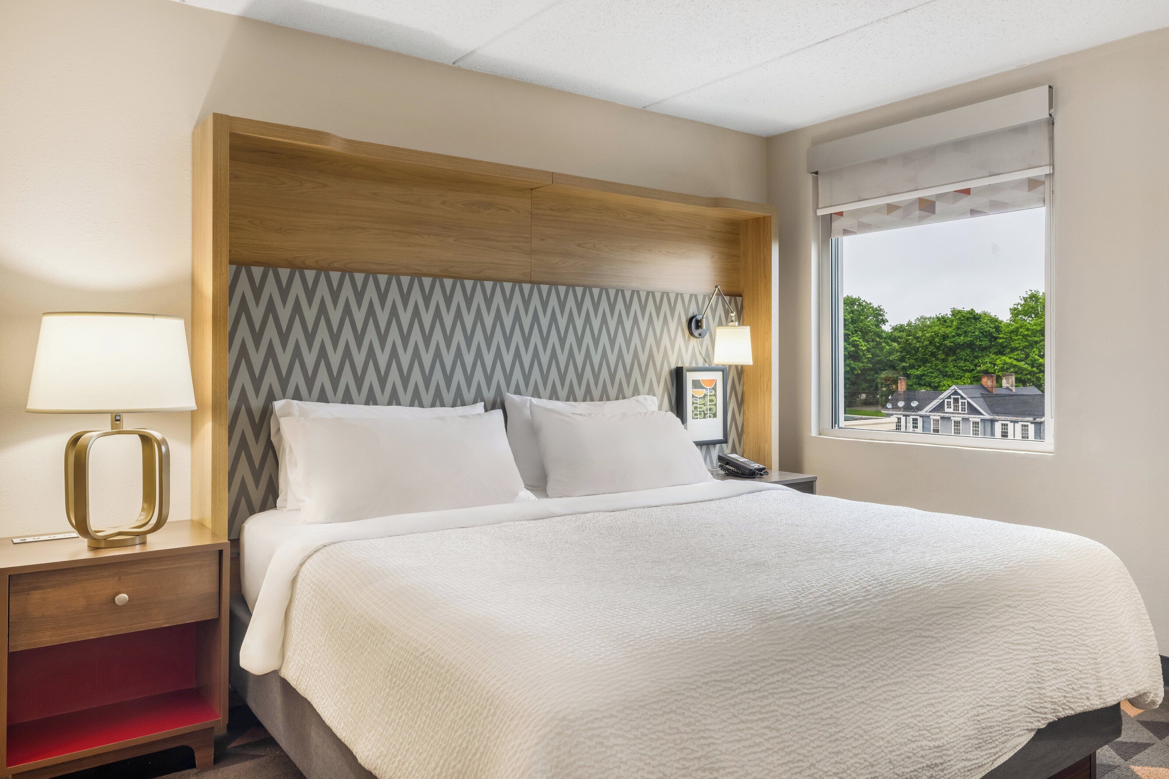A hotel room with a bed, bedside lamps, a nightstand, a window with a view, and a patterned wall.