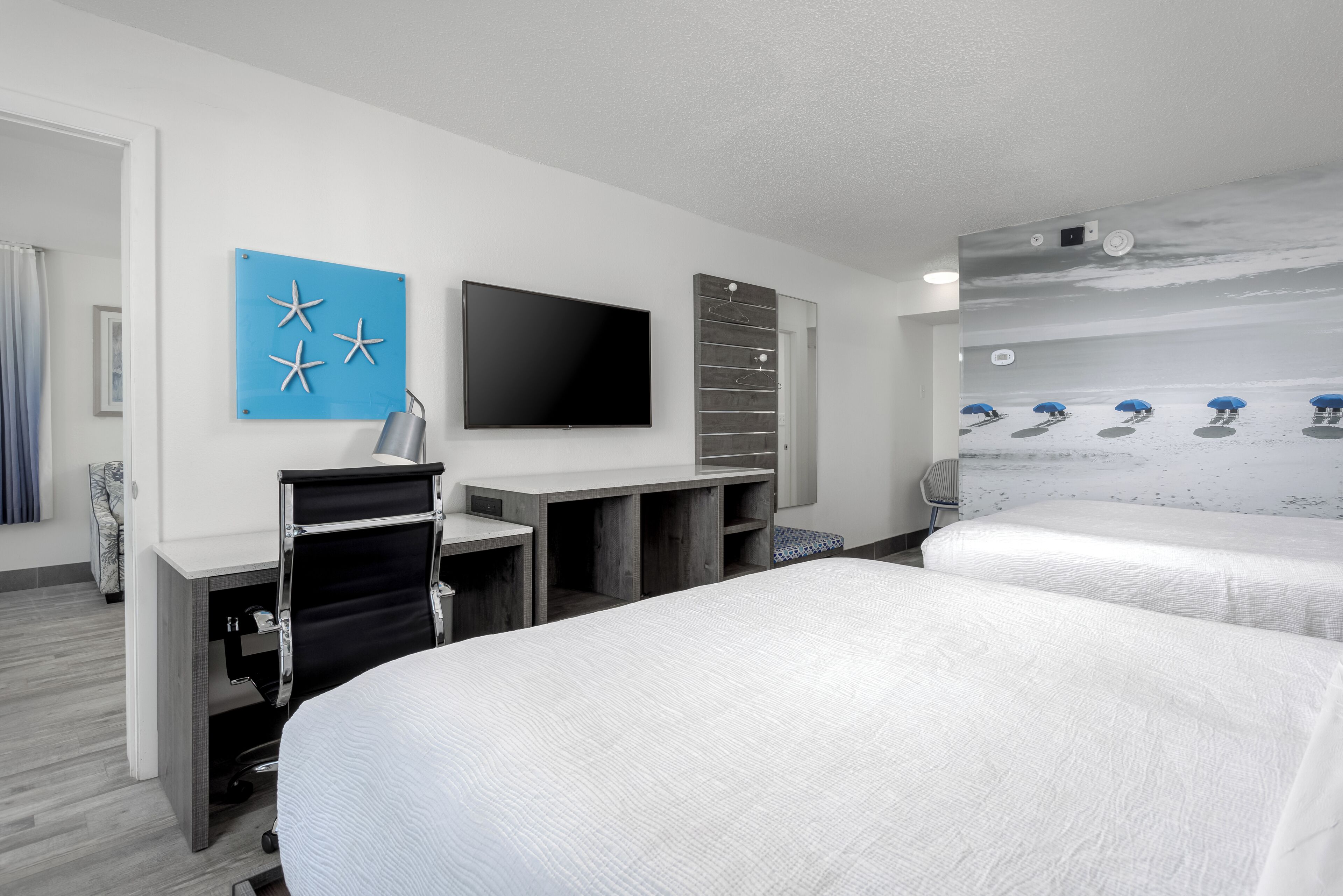 A hotel room with a flat-screen TV, a blue wall decoration, a wooden headboard, and a beach-themed wall mural.
