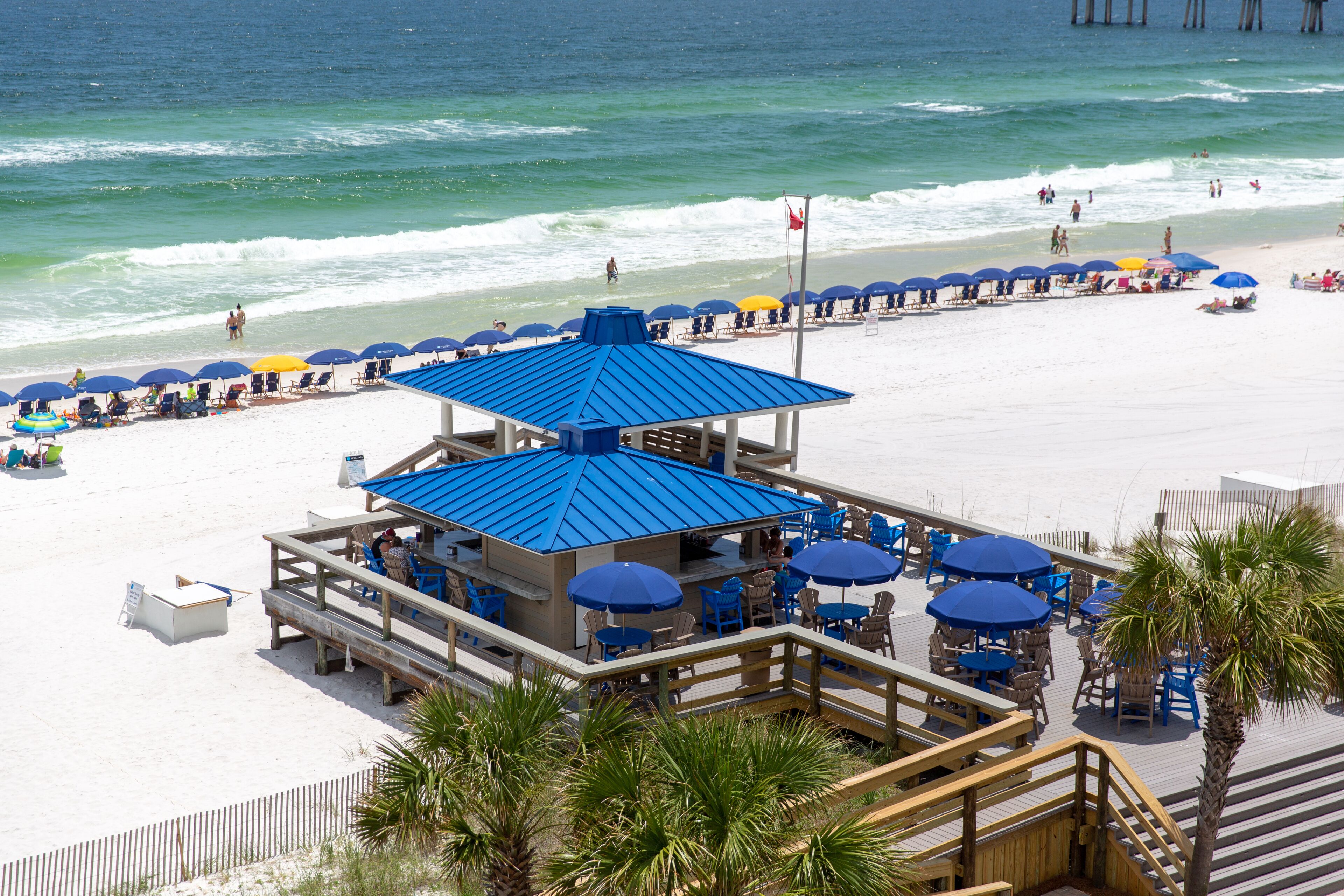 A beachside restaurant with outdoor seating and a blue roof, overlooking the ocean.