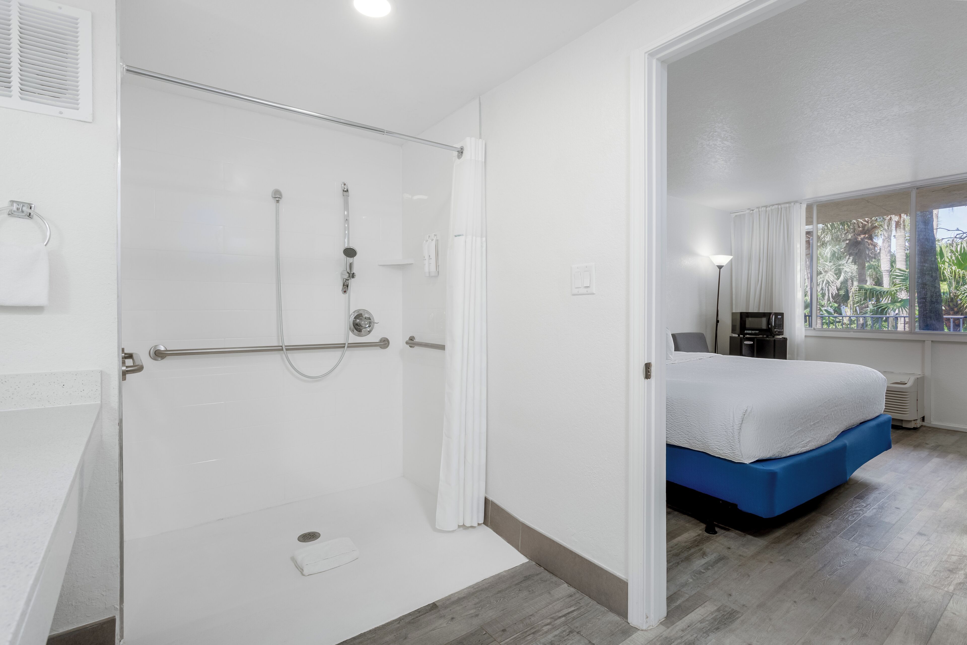 A hotel room with a shower, a bed, a window with a view of palm trees, and a bedside table.