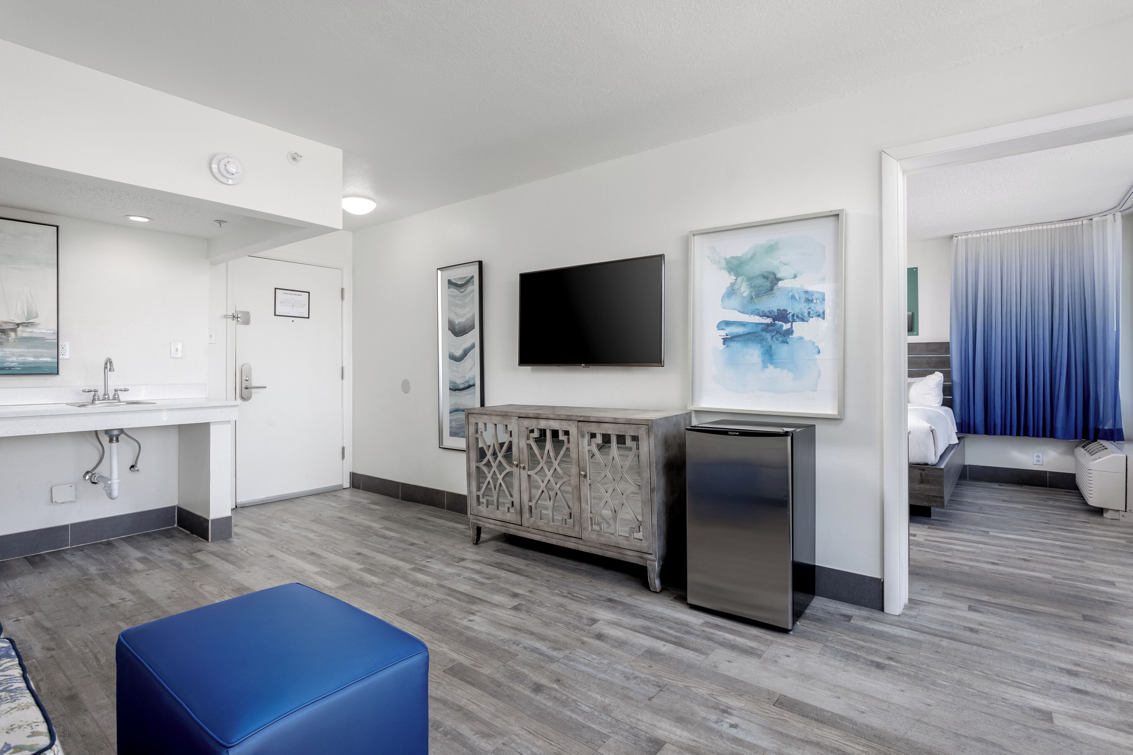 Habitación de hotel con kitchenette, televisión de pantalla plana, mueble con paneles decorativos y un otomano azul.