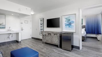Habitación de hotel con kitchenette, televisión de pantalla plana, mueble con paneles decorativos y un otomano azul.