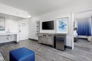 Habitación de hotel con kitchenette, televisión de pantalla plana, mueble con paneles decorativos y un otomano azul.