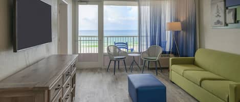Ein Hotelzimmer mit einem Holzkoffer, einem grünen Sofa, einem blauen Hocker, zwei Stühlen, einem kleinen Tisch und einem großen Fenster mit Blick auf den Ozean.