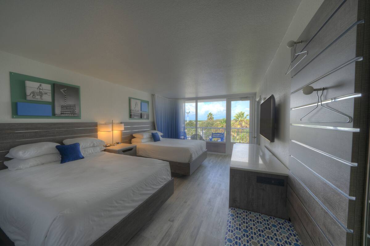 A hotel room with two beds, a desk, a TV, and a balcony with a view of palm trees.