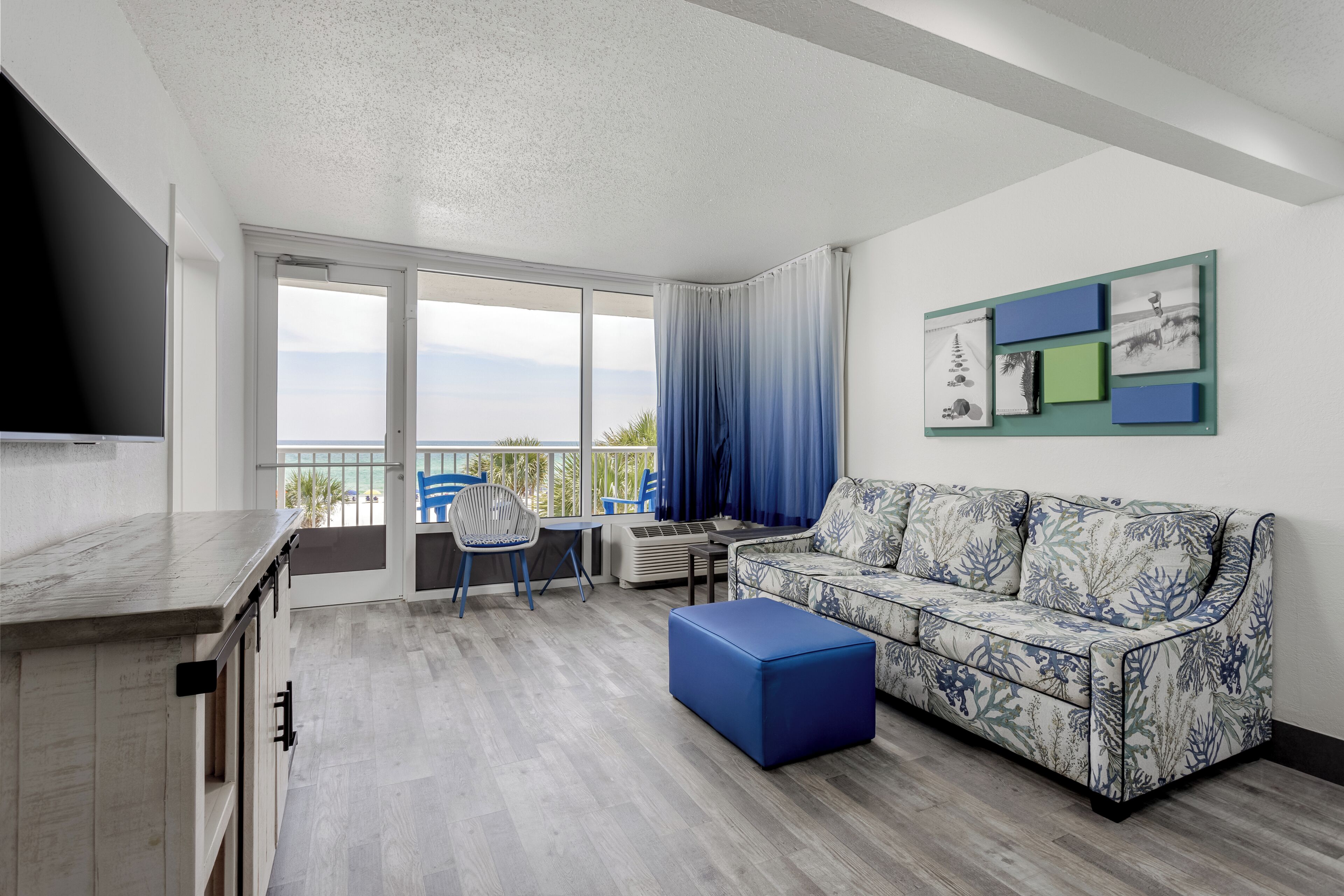 A modern living room with a sofa, a blue ottoman, a dining table with chairs, and a TV mounted on the wall.