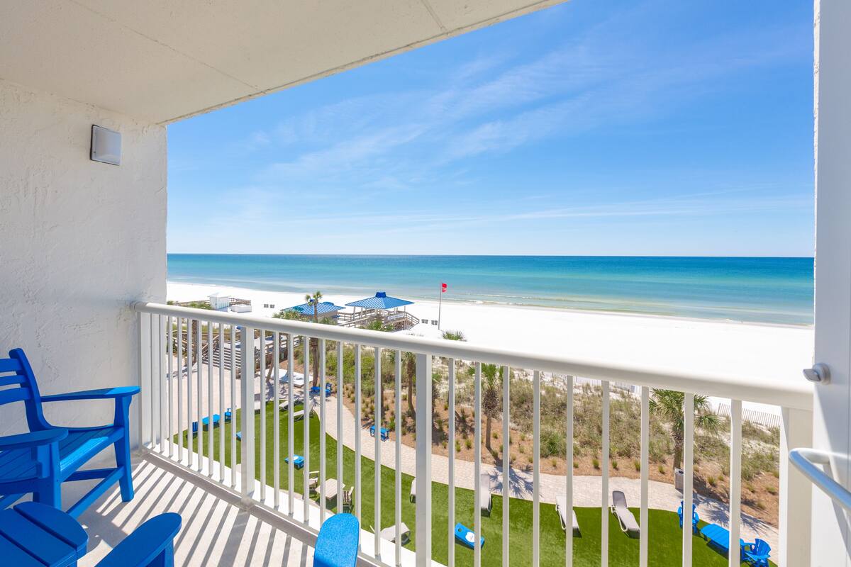 A balcony with a view of the beach, white railing, and blue chairs.