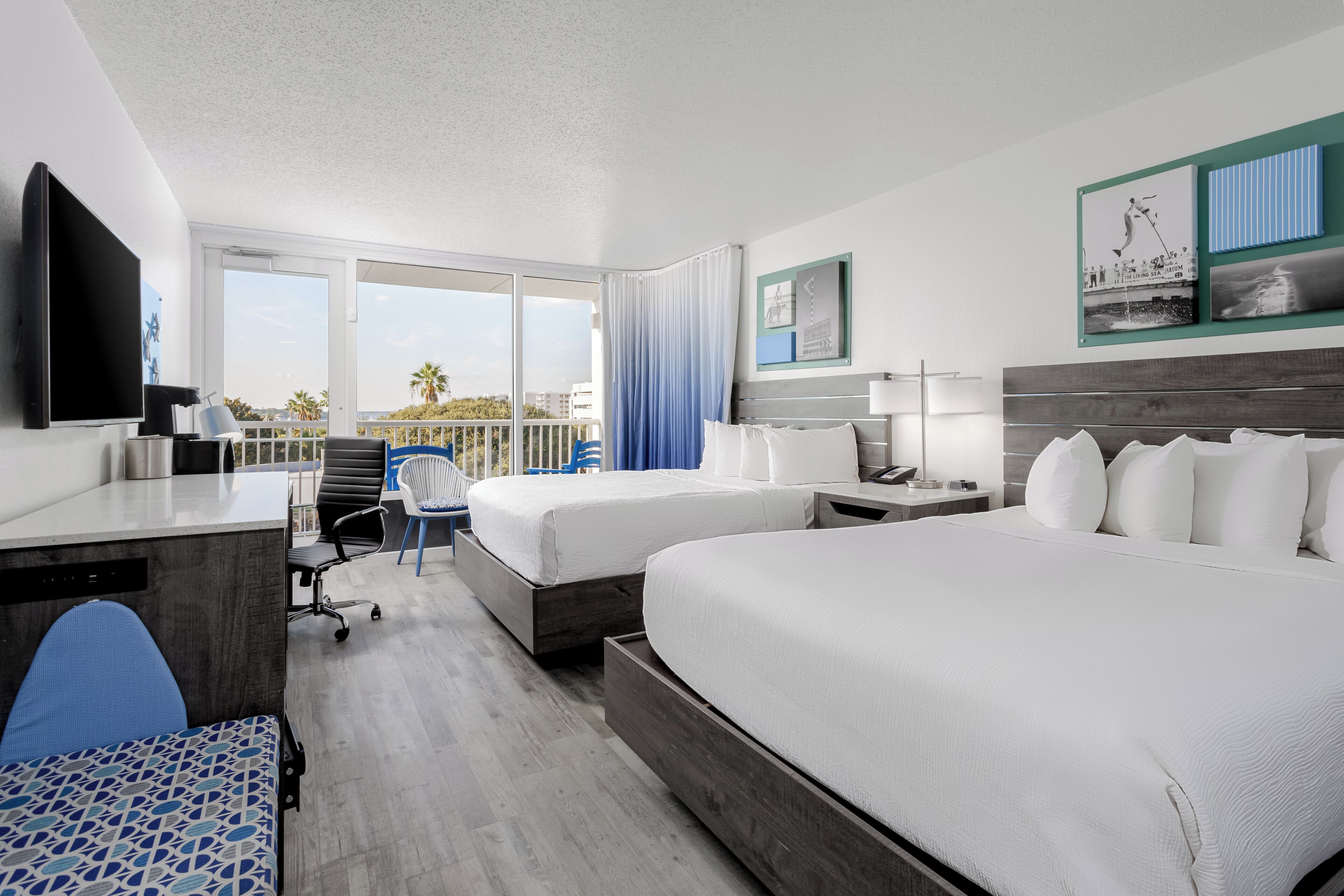 A hotel room with two beds, a desk, a chair, a TV, and a balcony with a view of palm trees and buildings.