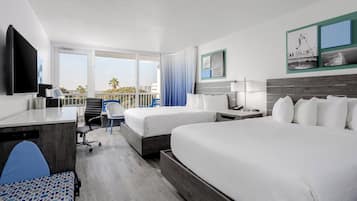 A hotel room with two beds, a desk, a chair, a TV, and a balcony with a view of palm trees and buildings.