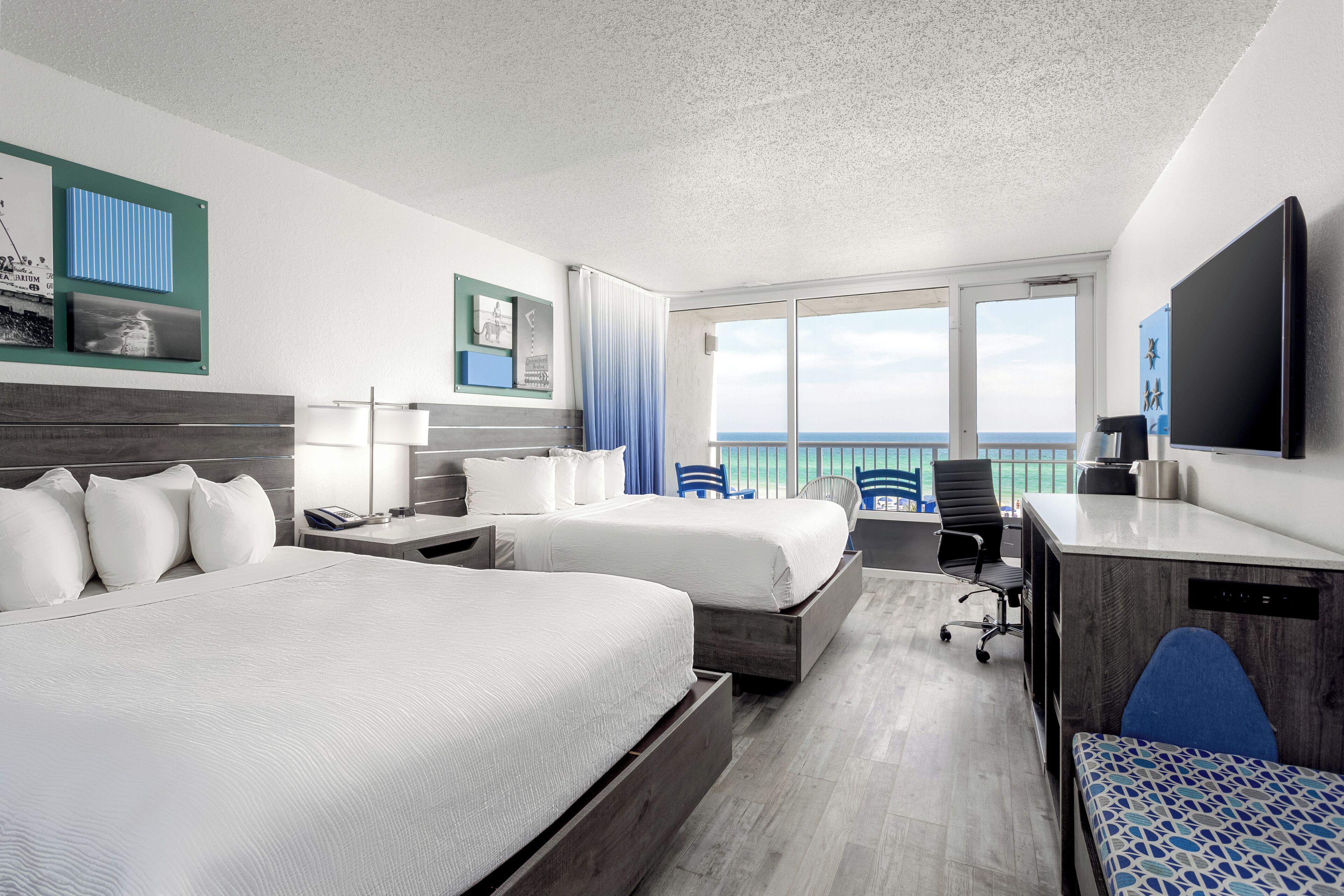 A hotel room with two beds, a desk, a chair, a TV, and a view of the ocean.