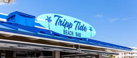 Un bar de plage appelé « Tripp Tide », avec un ciel bleu azur et des palmiers en arrière-plan.