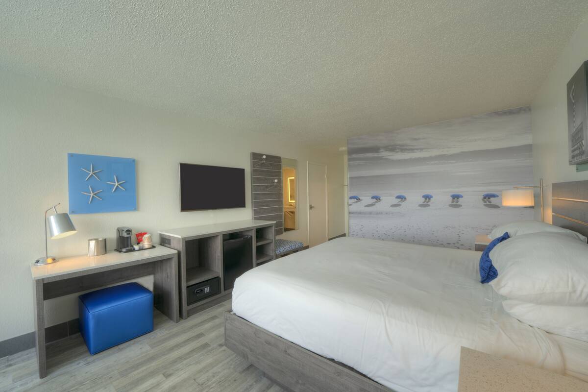 A hotel room with a bed, a desk, a TV, a wardrobe, and a wall with a beach mural.