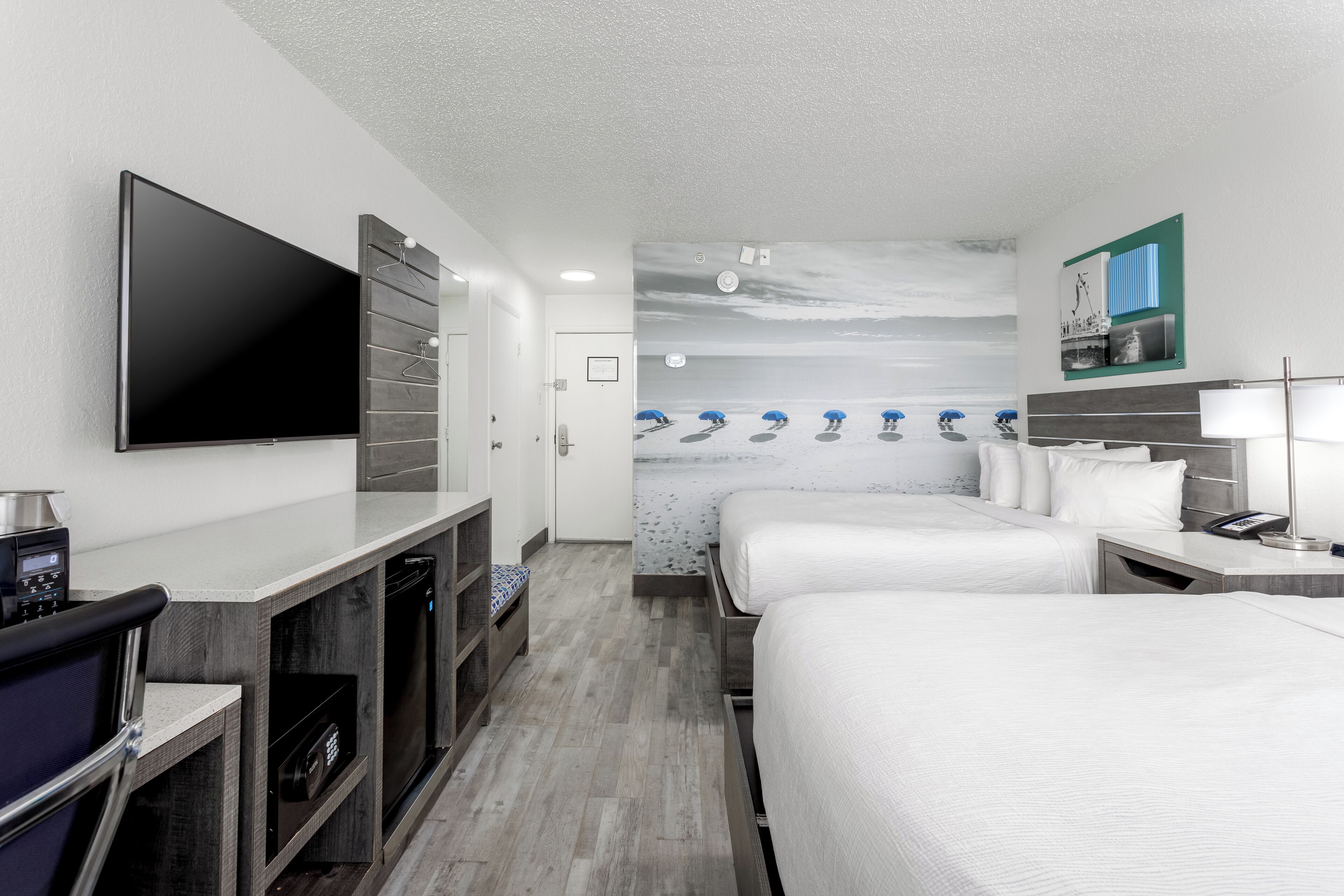 A hotel room with a flat-screen TV, a minibar, a desk with a coffee machine, two beds with white linens, a bathroom with a shower, and a wall with a beach mural.