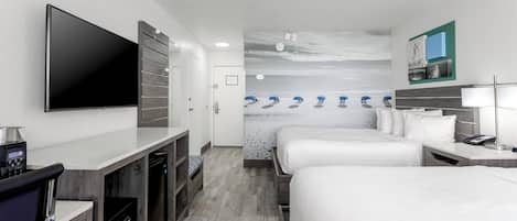 A hotel room with a flat-screen TV, a minibar, a desk with a coffee machine, two beds with white linens, a bathroom with a shower, and a wall with a beach mural.