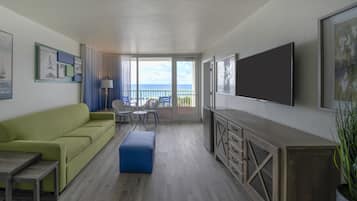 Habitación de hotel con sofá verde, otomana azul, televisor montado en la pared y balcón con vista al océano.