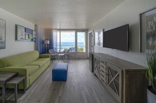 Habitación de hotel con sofá verde, otomana azul, televisor montado en la pared y balcón con vista al océano.