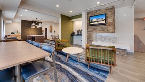 A modern hotel lobby with a long dining table, comfortable seating, a fireplace, and a television mounted on a stone wall. - Fairfield Inn & Suites by Marriott Spearfish (Spearfish)
