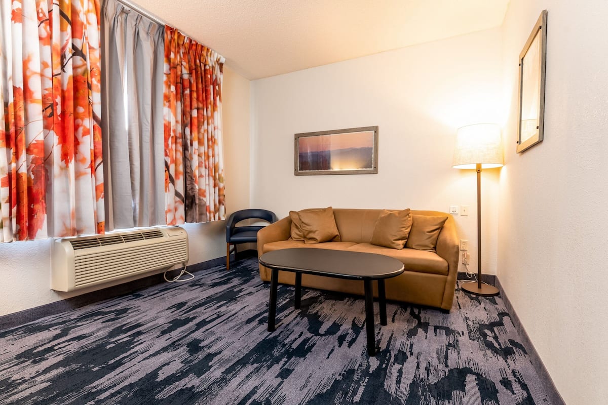 A hotel room with a sofa, a small table, a floor lamp, a radiator, and a window with curtains.