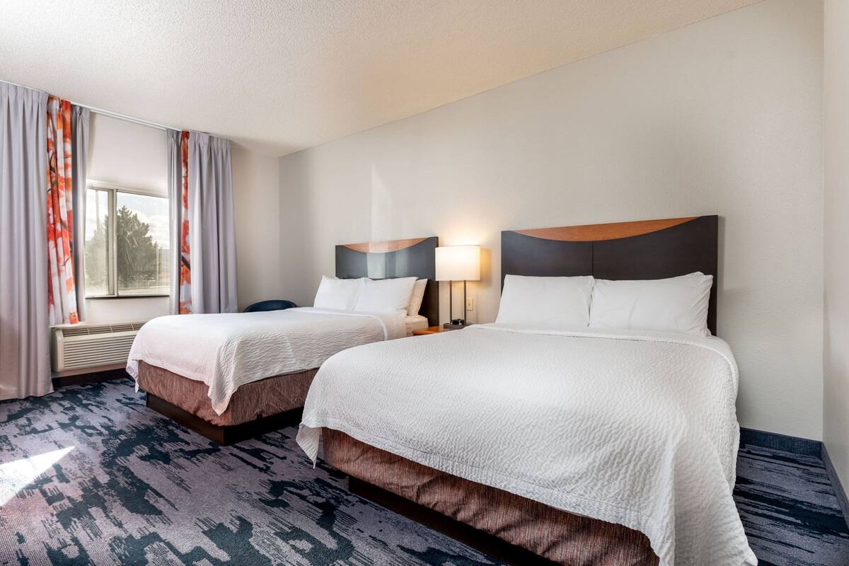 A hotel room with two beds, a window with curtains, a wall-mounted lamp, and a carpeted floor.