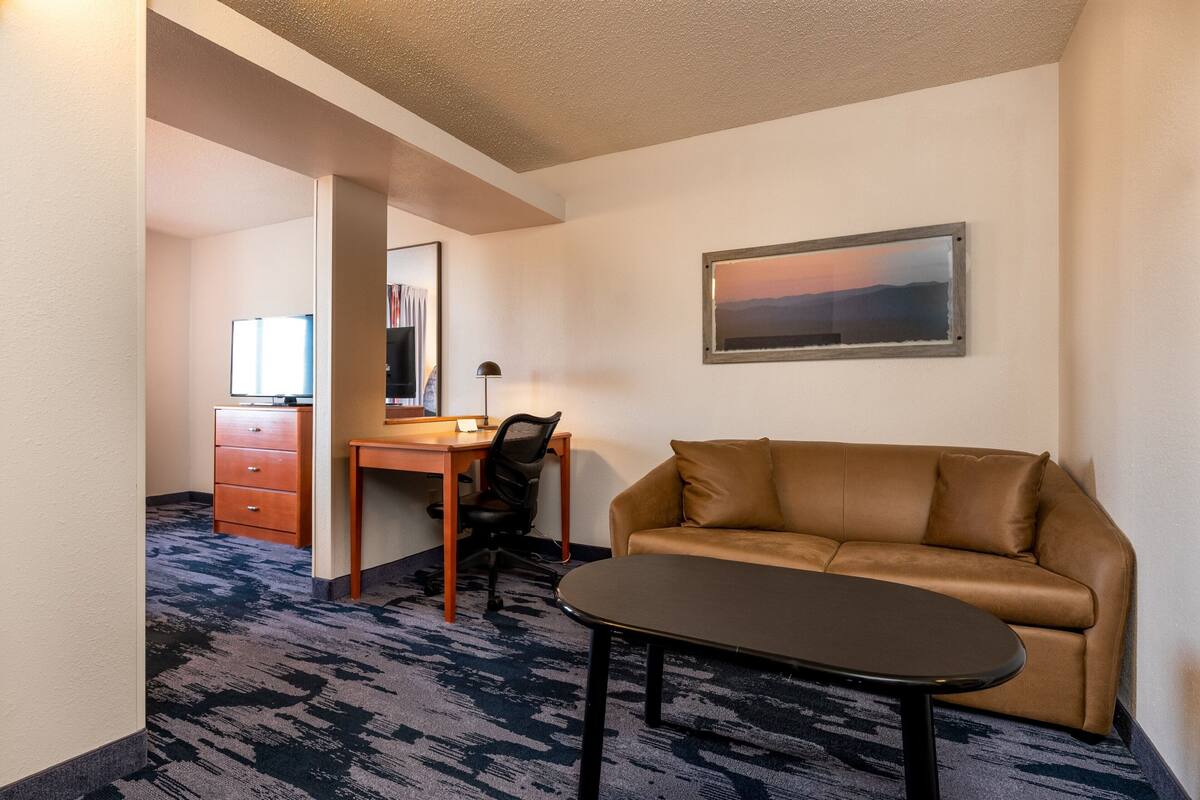 A hotel room with a brown leather sofa, a small table, a desk with a chair, and a framed picture on the wall.