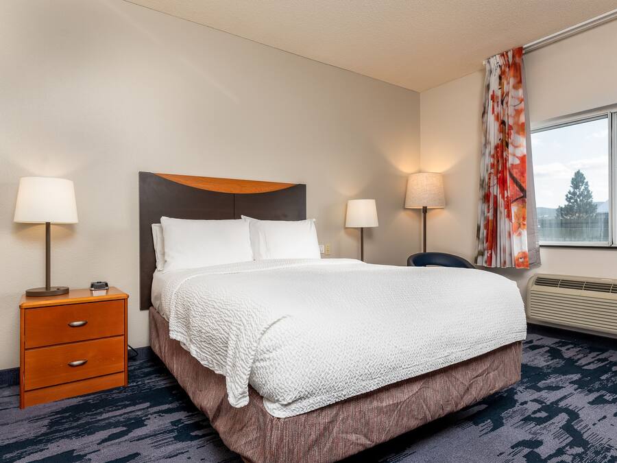 A hotel room with a bed, two bedside tables with lamps, a window with curtains, and a wall-mounted heater.