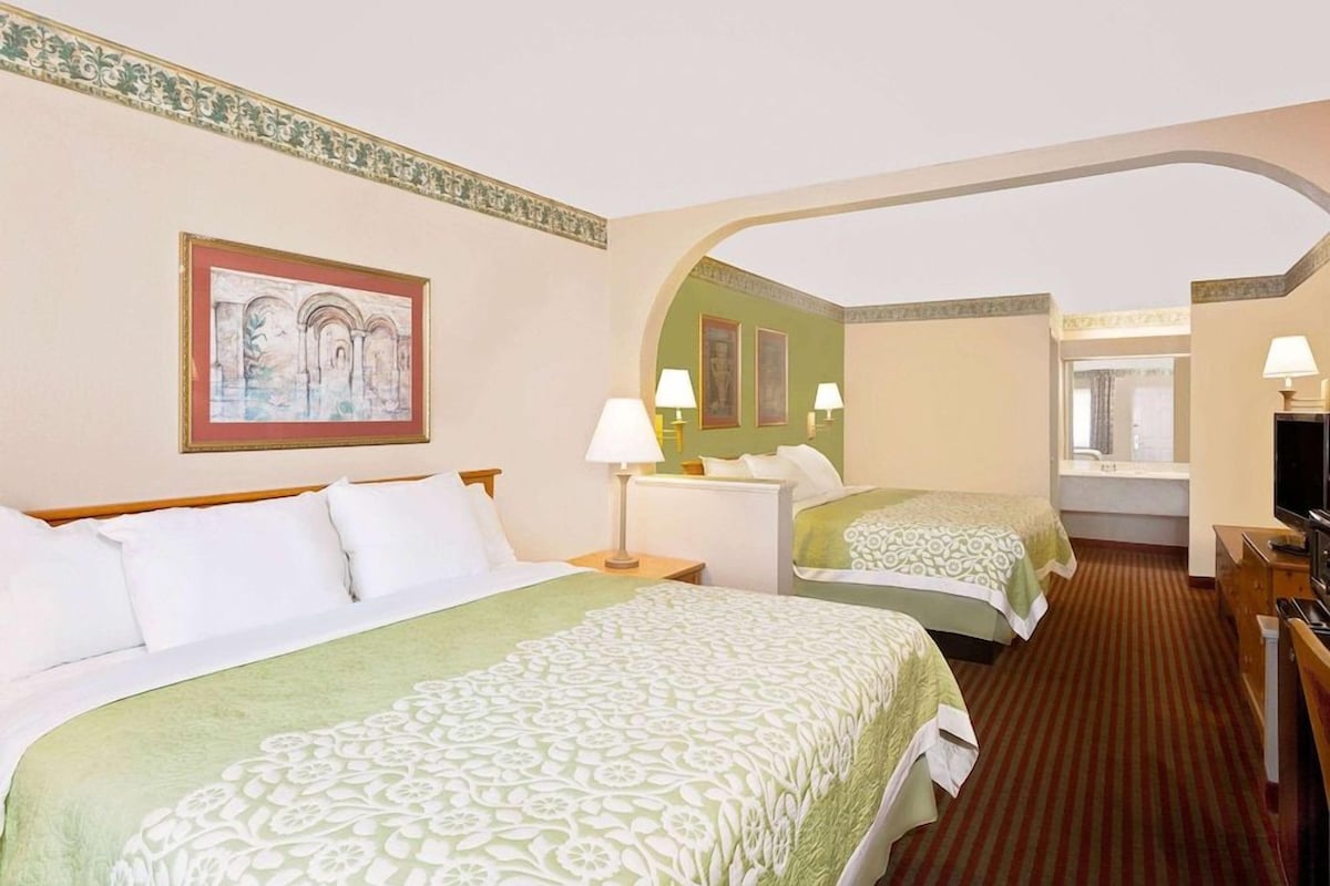 A hotel room with two beds, a television, a desk, and a bathroom visible through an archway.
