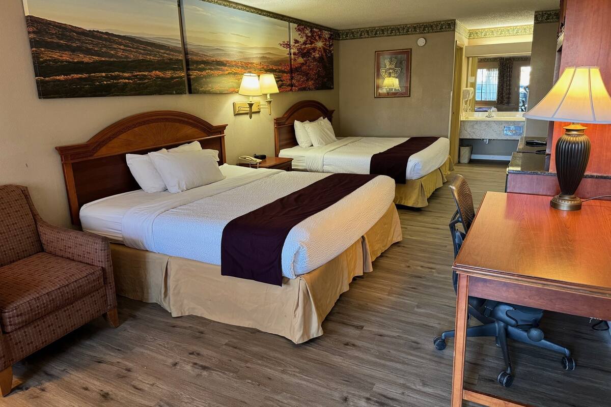 A hotel room with two beds, a desk, a chair, a lamp, a painting, and a mirror.