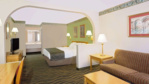Quarto de hotel com cama, sofá, escrivaninha, televisão e banheiro visível por um arco.