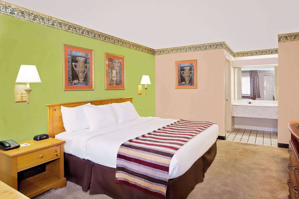 A hotel room with a bed, nightstand, lamp, and a bathroom visible in the background.