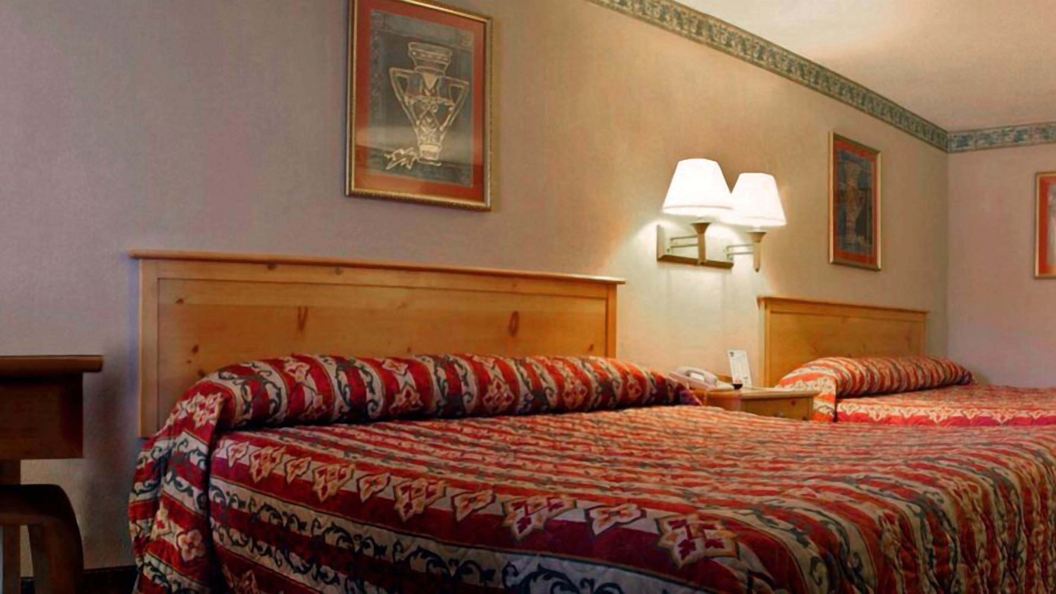A hotel room with two beds, wooden headboards, patterned bedspreads, a framed picture on the wall, and a wall-mounted lamp.
