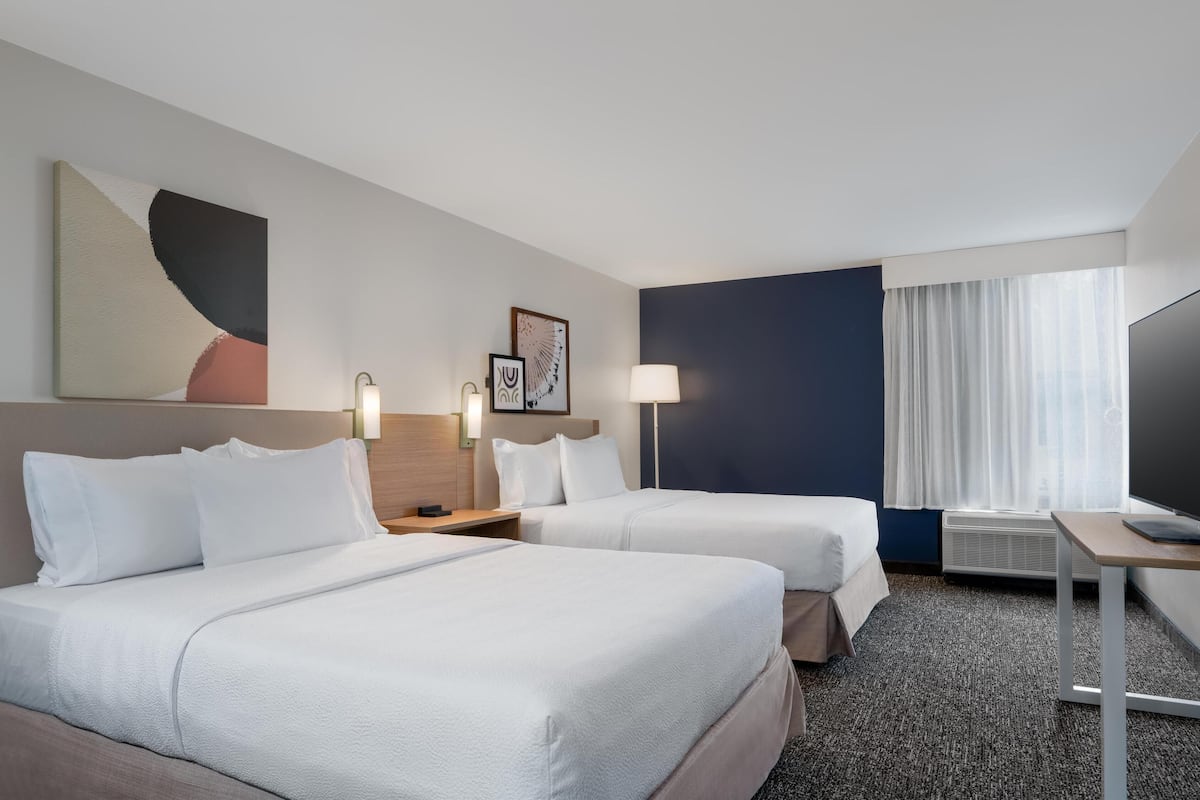 A hotel room with two beds, a desk, a television, and a large window with curtains.