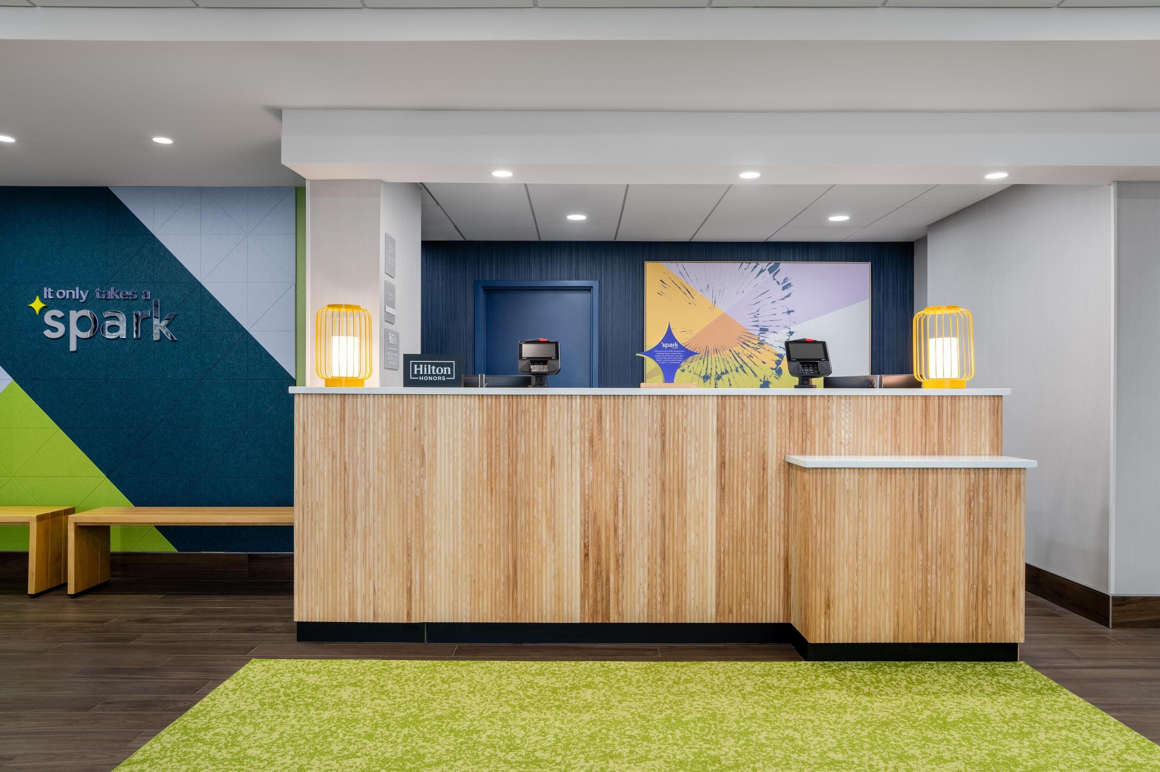 En receptionsdisk med träpanel, två vägglampor, en blå vägg med en skylt som visar "Hilton Honors", och en grön, mönstrad matta.