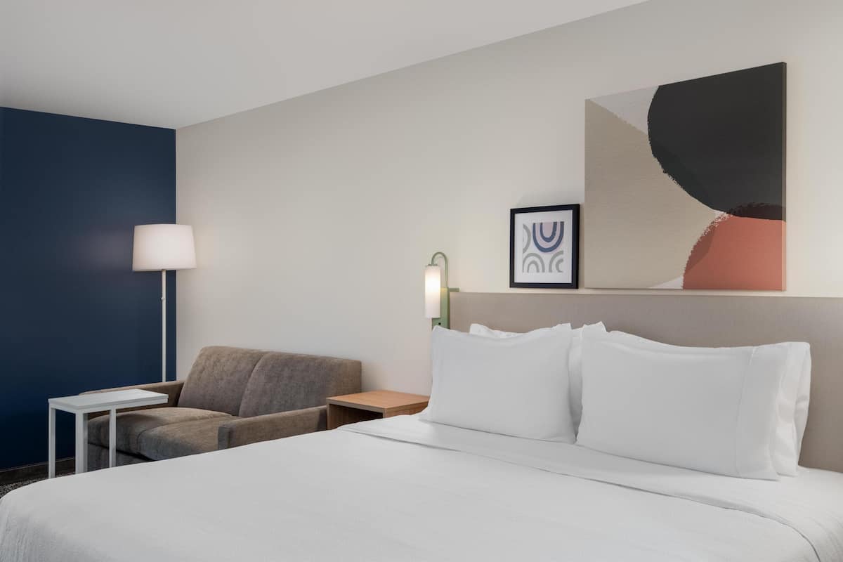 A hotel room with a bed, a desk, a chair, a lamp, and a wall art piece.