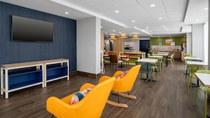 A modern dining area with wooden tables and chairs, a flat-screen TV mounted on the wall, and a kitchen area in the background.
