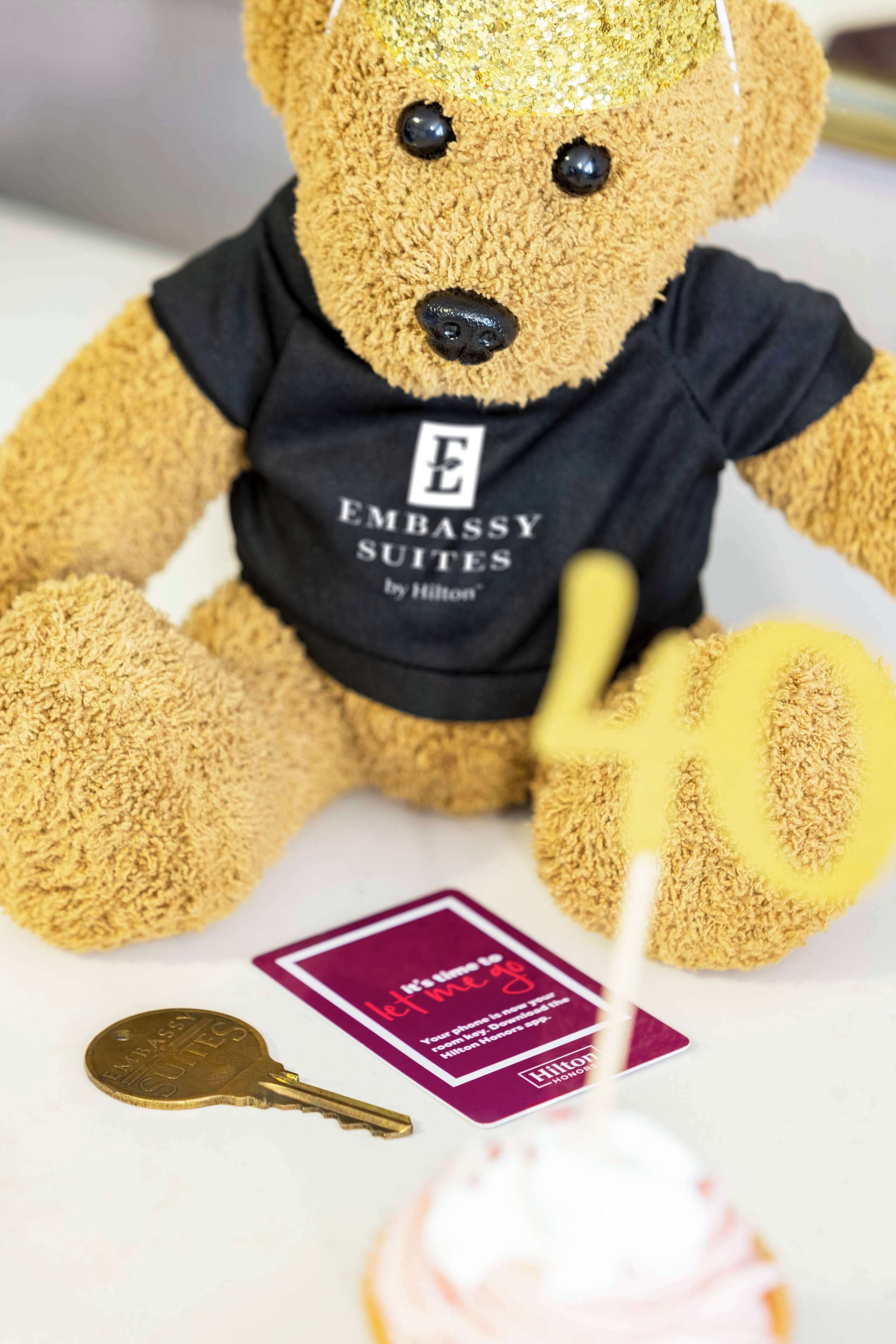 A plush teddy bear wearing a black t-shirt with "EMBASSY SUITES by Hilton" printed on it, sitting on a table with a golden key and a pink cake with a candle.