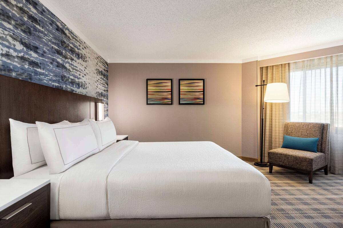 Ein Hotelzimmer mit einem großen Bett, einem Sessel, einem Schreibtisch und zwei gerahmten Kunstwerken an der Wand.